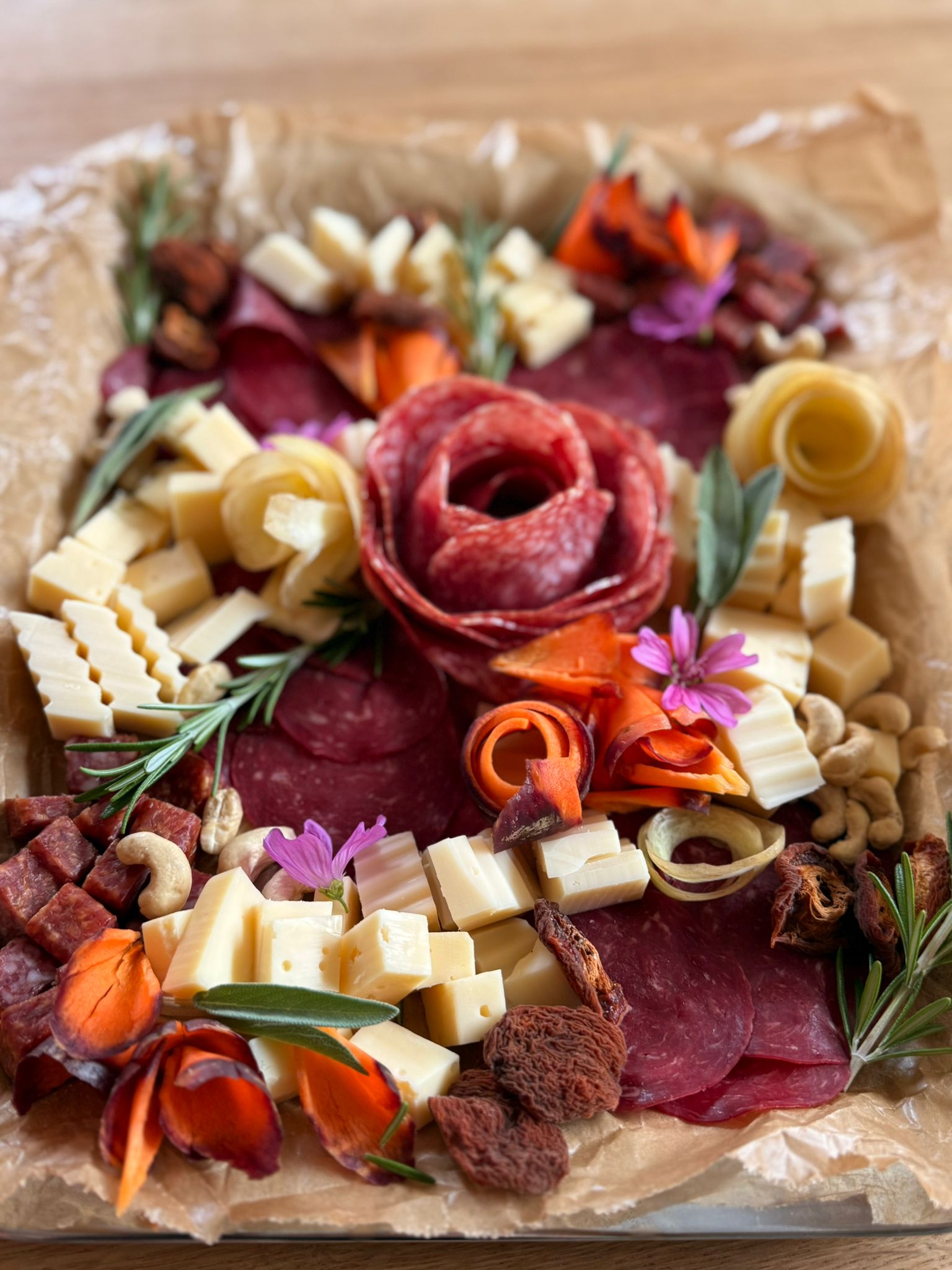 Charcuterie-Platte mit Käsewürfeln, Salami in Rosenform, Gemüseblüten, Nüssen und Kräutern.